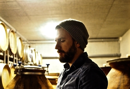 Mann mit Mütze verkostet Wein im Weinkeller, umgeben von Amphoren.