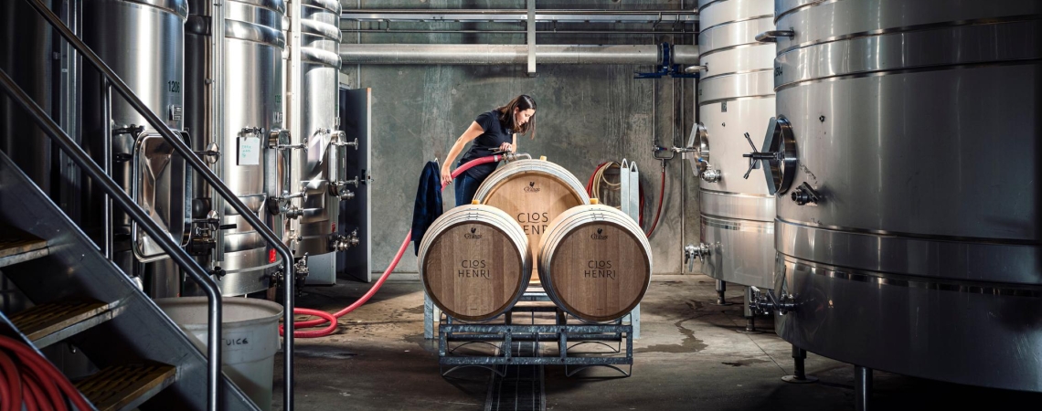 Frau arbeitet in einer modernen Weinkellerei mit großen Edelstahltanks und Holzfässern.