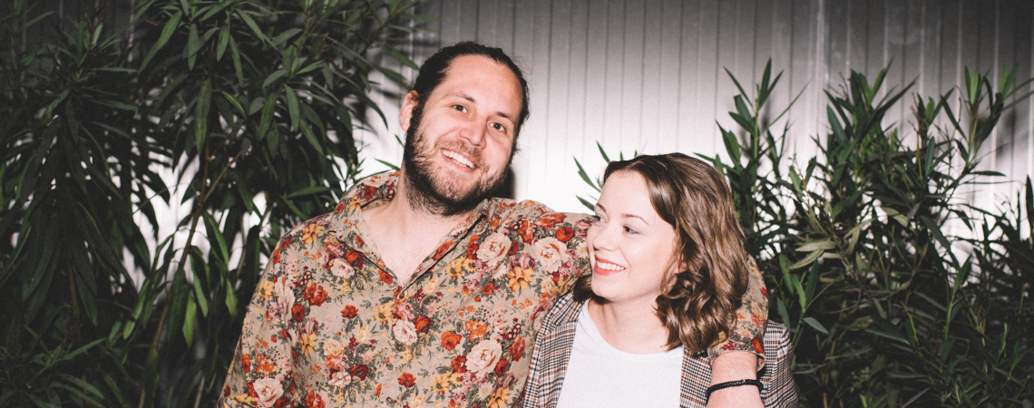 Zwei lächelnde Personen vor Pflanzen, eine trägt ein florales Hemd, die andere karierten Blazer.