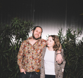 Zwei lächelnde Personen vor Pflanzen, eine trägt ein florales Hemd, die andere karierten Blazer.