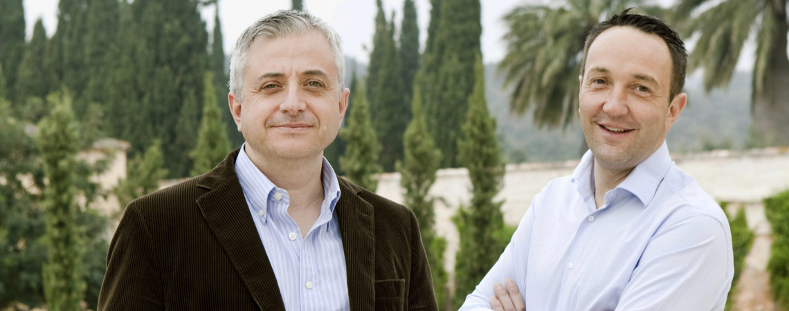 Zwei Männer in Freizeitkleidung vor bewaldeter Hintergrundleandschaft.