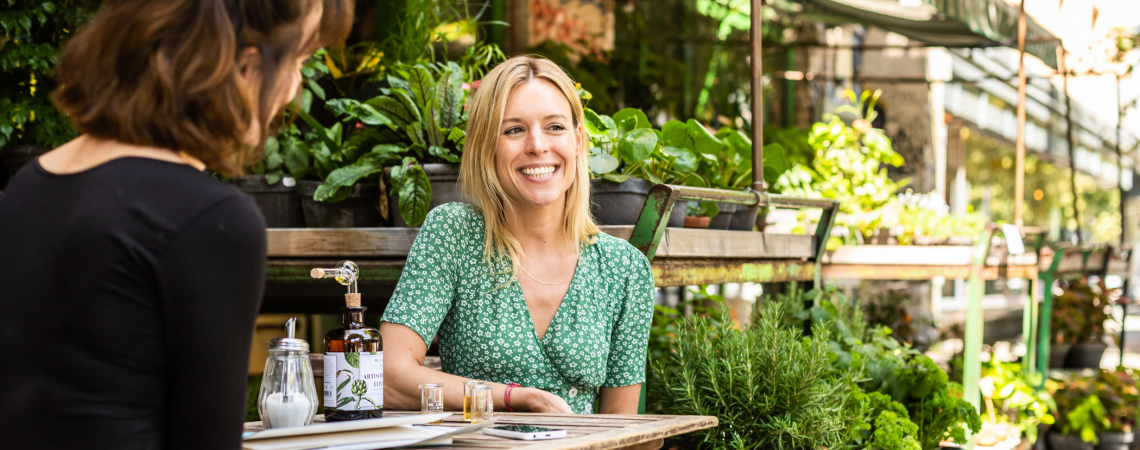 Zwei Frauen sitzen lächelnd im grünen, gemütlichen Straßencafé mit Pflanzen.