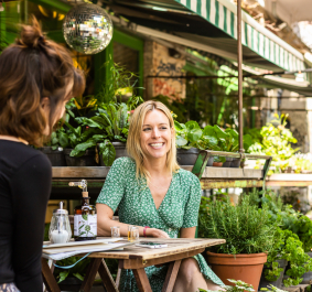 Zwei Frauen sitzen lächelnd im grünen, gemütlichen Straßencafé mit Pflanzen.