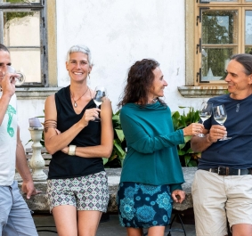 Vier Personen stehen draußen lachend mit Weingläsern, umgeben von Pflanzen und Steinmöbeln.