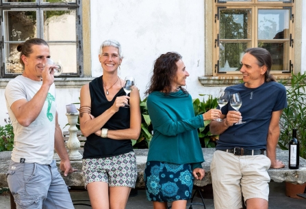 Vier Personen stehen draußen lachend mit Weingläsern, umgeben von Pflanzen und Steinmöbeln.