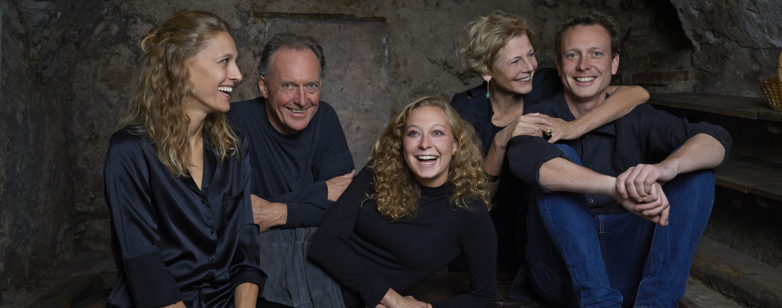 Fünf lachende Personen in schwarzer Kleidung, sitzend auf einer rustikalen Holzbank.