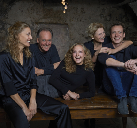 Fünf lachende Personen in schwarzer Kleidung, sitzend auf einer rustikalen Holzbank.