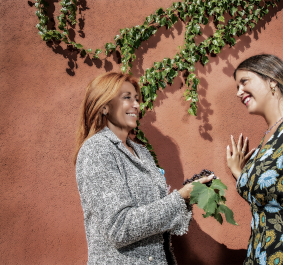 Zwei Frauen lächelnd vor bemooster Terracotta-Mauer mit Weinranken.
