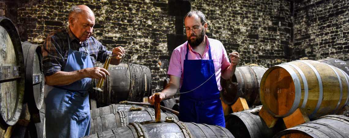 Zwei Männer entnehmen Proben aus Holzfässern in einem Gewölbekeller.