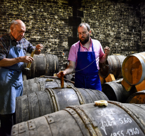 Zwei Männer entnehmen Proben aus Holzfässern in einem Gewölbekeller.