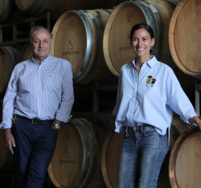 Zwei Personen posieren vor gestapelten Weinfässern in einem Weinkeller.