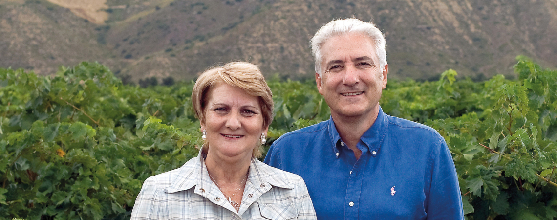Zwei Menschen stehen lächelnd in einem grünen Weinberg vor hügeliger Landschaft.