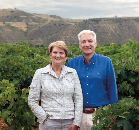 Zwei Menschen stehen lächelnd in einem grünen Weinberg vor hügeliger Landschaft.