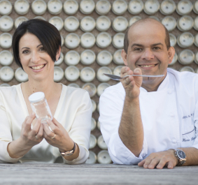 Zwei Personen lächeln, halten Glas und Gabel vor gemusterter Hintergrundwand aus Metallscheiben.
