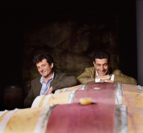 Zwei Männer lehnen an alten Weinfässern in einem dunklen Keller.