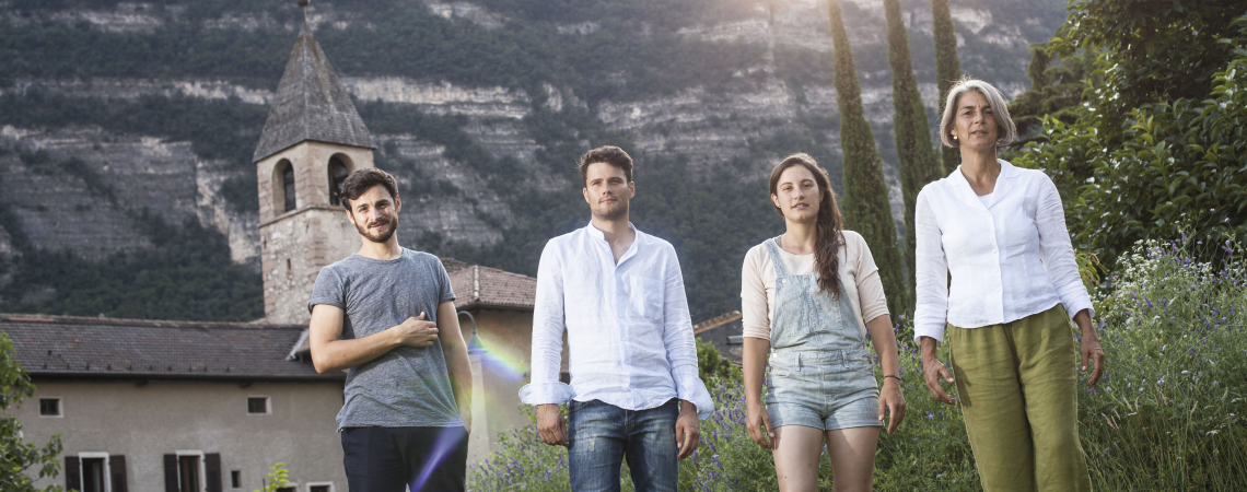 Vier Personen vor ländlicher Kulisse mit Berg und Kirchturm im Hintergrund.