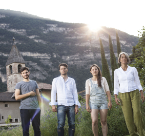 Vier Personen vor ländlicher Kulisse mit Berg und Kirchturm im Hintergrund.