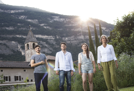 Vier Personen vor ländlicher Kulisse mit Berg und Kirchturm im Hintergrund.