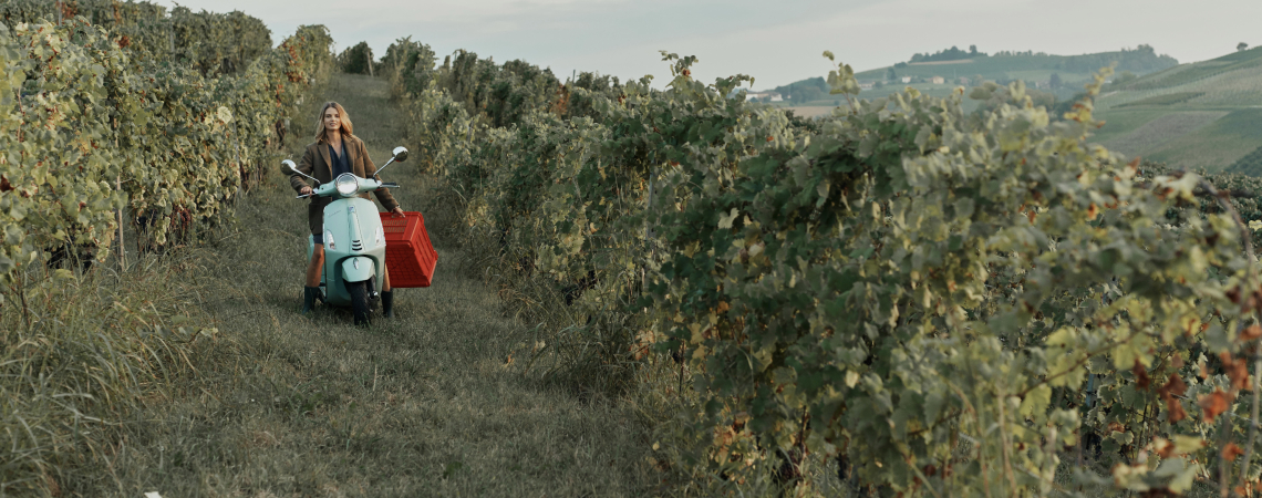 Frau auf hellblauem Roller in einem grünen Weinberg, dahinter hügelige Landschaft.