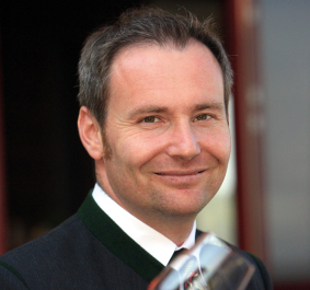 Mann in traditioneller Kleidung mit Glas Rotwein, lächelnd in die Kamera.