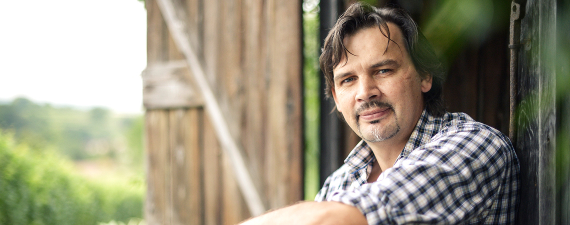 Mann in kariertem Hemd sitzt entspannt vor Holzhütte im Grünen.