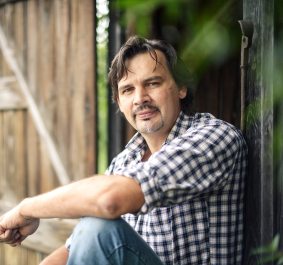 Mann in kariertem Hemd sitzt entspannt vor Holzhütte im Grünen.