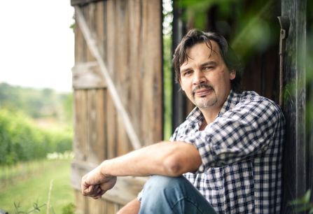 Mann in kariertem Hemd sitzt entspannt vor Holzhütte im Grünen.