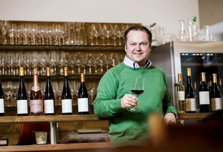 Mann in grünem Pullover hält Weinglas, umgeben von Weinflaschen und Gläsern.