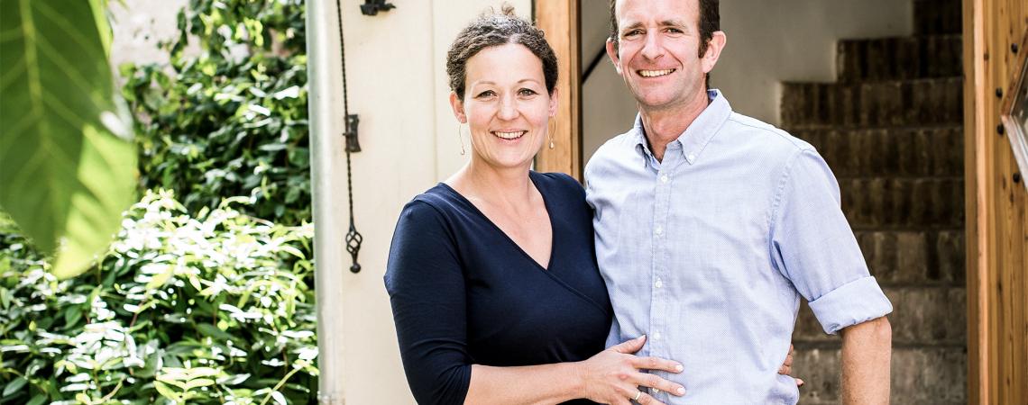 Paar lächelnd vor steinverzierter Tür, umgeben von Pflanzen.