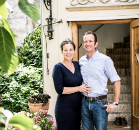 Paar lächelnd vor steinverzierter Tür, umgeben von Pflanzen.