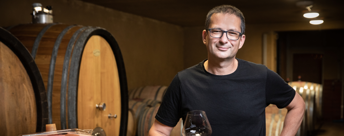 Mann in Weinkeller mit großen Holzfässern, hält ein Glas Rotwein.