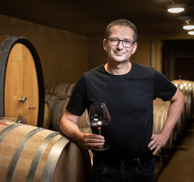 Mann in Weinkeller mit großen Holzfässern, hält ein Glas Rotwein.