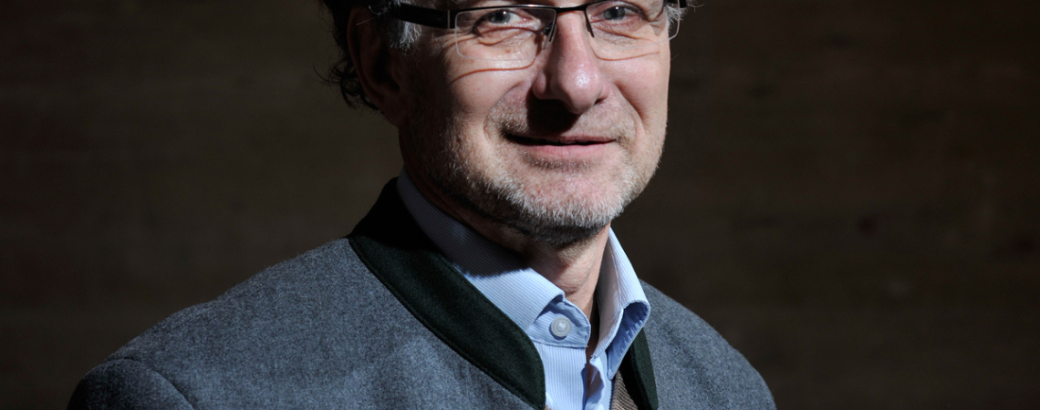 Mann im Trachtenjanker mit Brille hält ein Glas Weißwein, dunkler Hintergrund.