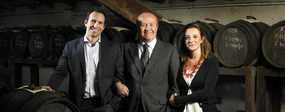 Drei Personen in eleganter Kleidung stehen lächelnd in einem Weinkeller vor Holzfässern.