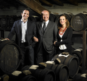 Drei Personen in eleganter Kleidung stehen lächelnd in einem Weinkeller vor Holzfässern.