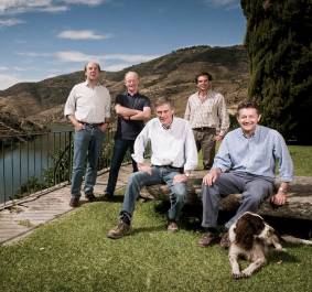 Fünf Männer und ein Hund posieren vor malerischer Flusslandschaft und Hügeln.