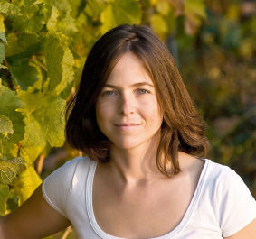 Frau in weißem T-Shirt steht neben grünen Weinreben im Sonnenlicht.