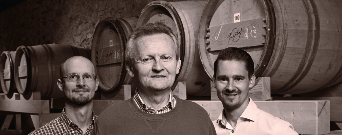 Drei Männer vor Holzfässern in einem Kellerraum, sepiafarbene Aufnahme.