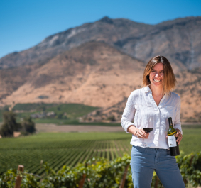 Frau in Weinberg mit Rotweinglas und Flasche vor Bergkulisse.