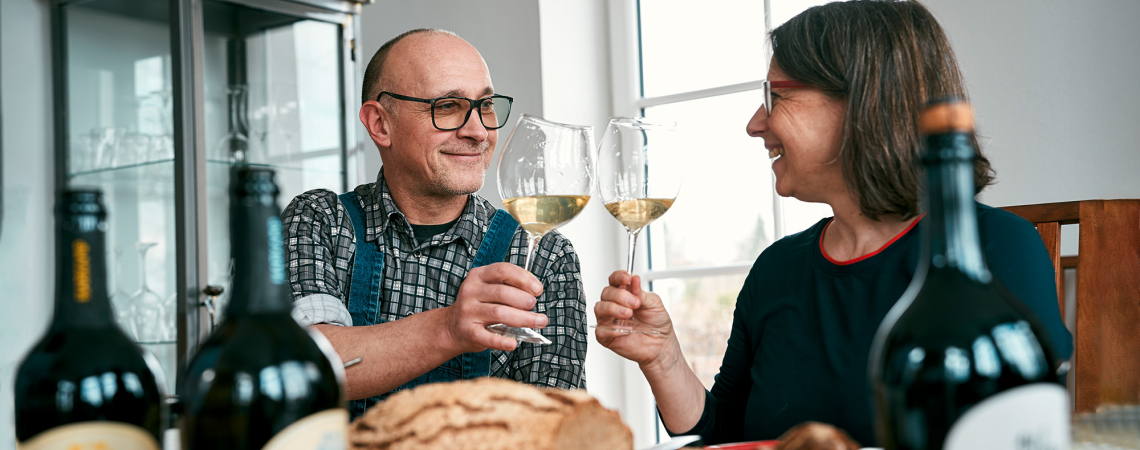 Zwei Personen stoßen lächelnd mit Weingläsern an, umgeben von Essen und Weinflaschen.