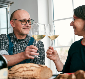 Zwei Personen stoßen lächelnd mit Weingläsern an, umgeben von Essen und Weinflaschen.