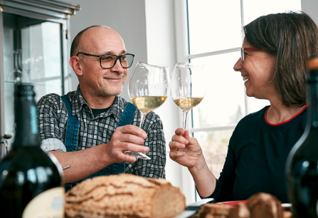 Zwei Personen stoßen lächelnd mit Weingläsern an, umgeben von Essen und Weinflaschen.