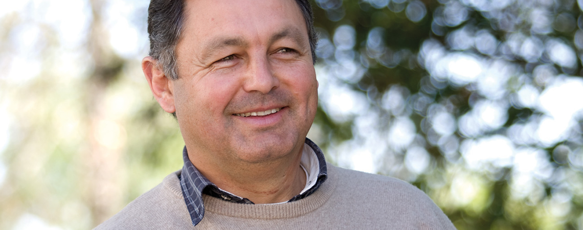 Mann in beigem Pullover hält ein Glas Weißwein, lächelnd im Freien.