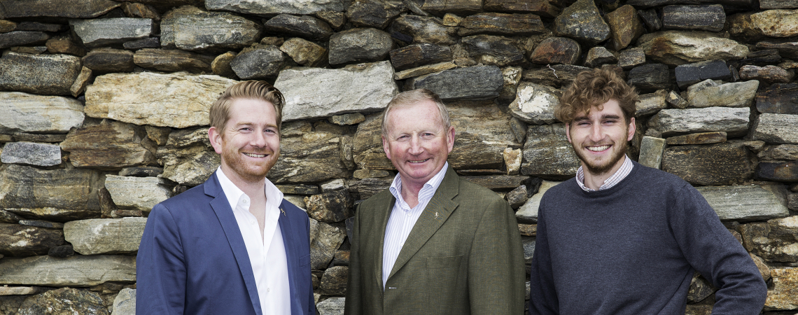 Drei Männer in Freizeitkleidung vor einer rustikalen Steinmauer.