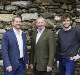 Drei Männer in Freizeitkleidung vor einer rustikalen Steinmauer.