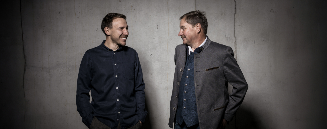 Zwei Männer in Freizeit- und Trachtenkleidung vor grauer Betonwand, lachen sich an.