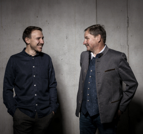 Zwei Männer in Freizeit- und Trachtenkleidung vor grauer Betonwand, lachen sich an.