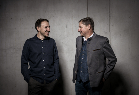 Zwei Männer in Freizeit- und Trachtenkleidung vor grauer Betonwand, lachen sich an.