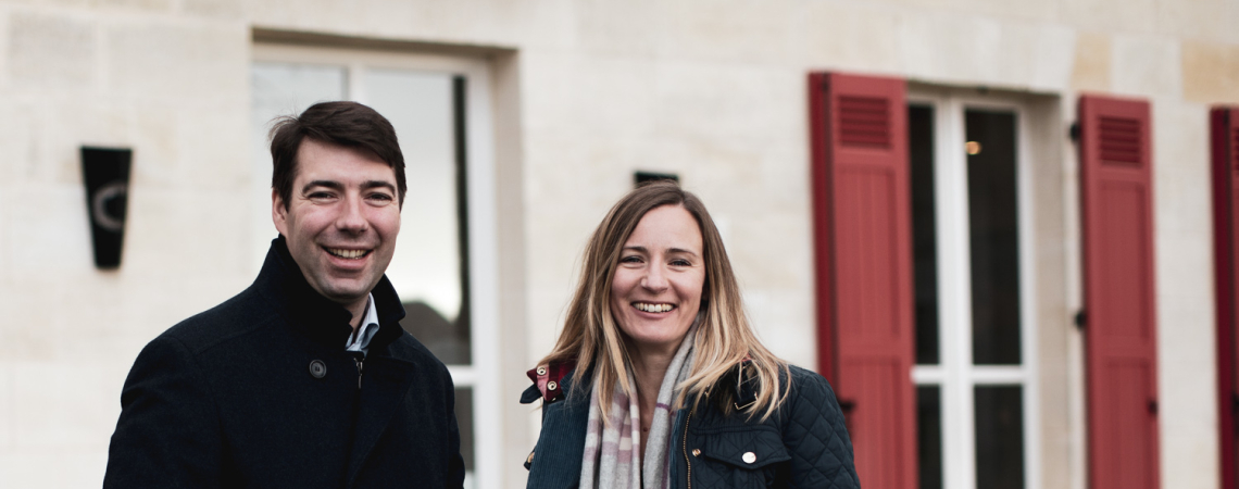 Zwei Personen in dunklen Mänteln vor einem Haus mit roten Fensterläden.