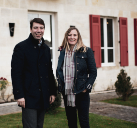 Zwei Personen in dunklen Mänteln vor einem Haus mit roten Fensterläden.